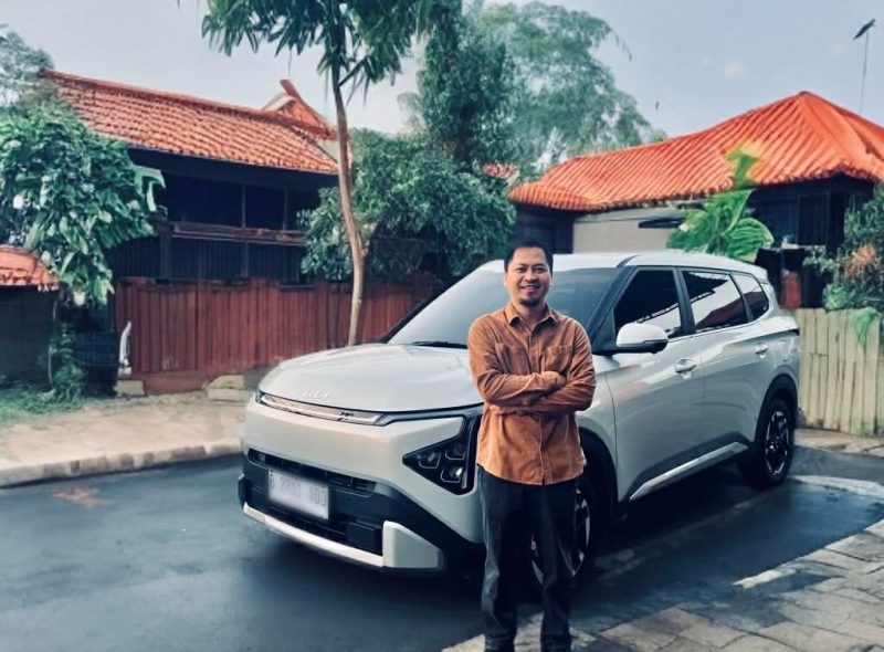 Seorang konsumen berpose di depan Kia Carens usai penggunaan harian dan perjalanan jarak menengah, menyoroti kenyamanan kabin serta fitur yang mendukung mobilitas keluarga.