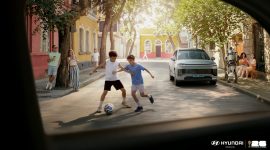 Dua anak bermain sepak bola di jalan lingkungan dengan latar mobil Hyundai Motor Company, sementara robot humanoid dari Boston Dynamics terlihat berinteraksi di sisi jalan, menggambarkan kolaborasi teknologi dan semangat FIFA World Cup 2026.