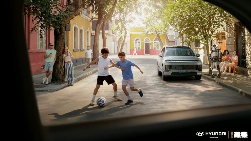 Dua anak bermain sepak bola di jalan lingkungan dengan latar mobil Hyundai Motor Company, sementara robot humanoid dari Boston Dynamics terlihat berinteraksi di sisi jalan, menggambarkan kolaborasi teknologi dan semangat FIFA World Cup 2026.