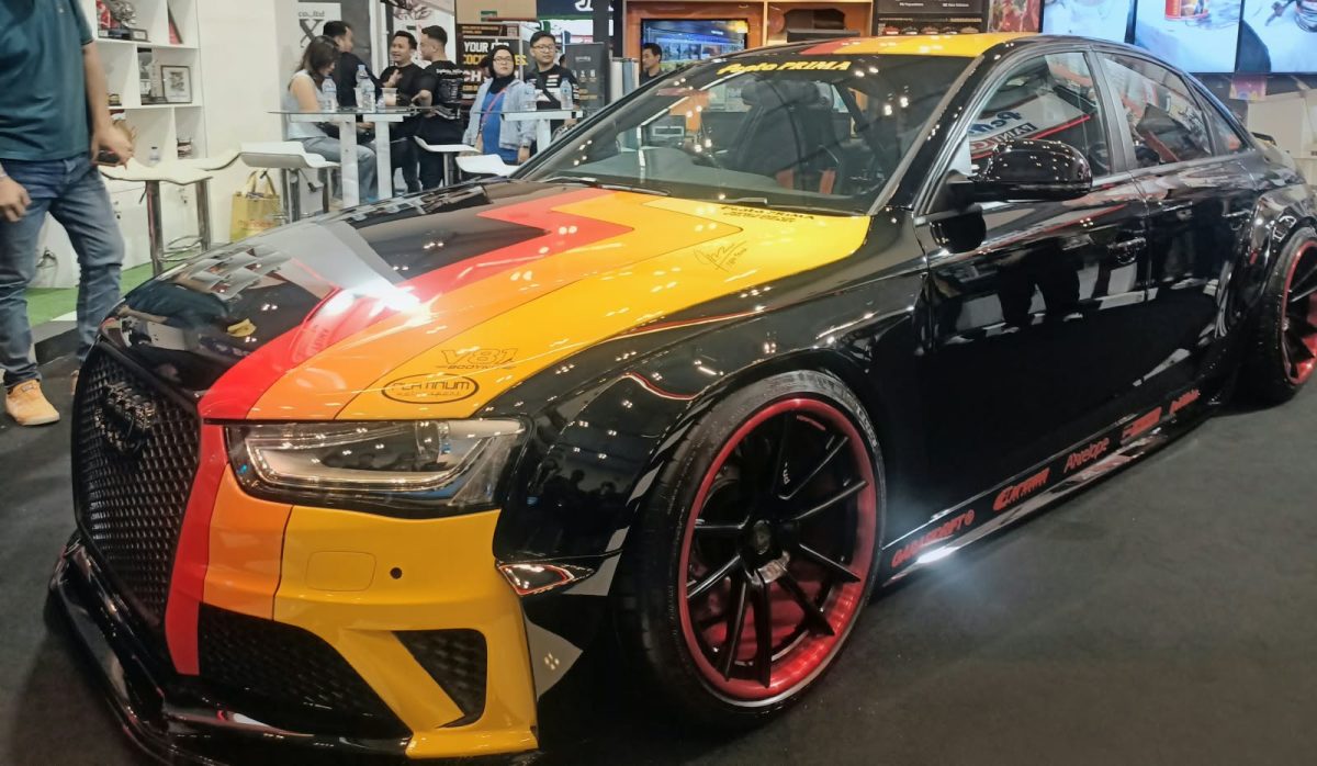 Keterangan Foto: Mobil modifikasi berbalut warna Obsidian Black dengan kombinasi grafis kuning, oranye, dan merah tampil di booth Penta Prima Paint pada ajang IMX 2026 di Tangerang. Finishing hitam mengilap dipadukan dengan velg sporty beraksen merah, memperkuat karakter agresif kendaraan.