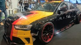 Keterangan Foto: Mobil modifikasi berbalut warna Obsidian Black dengan kombinasi grafis kuning, oranye, dan merah tampil di booth Penta Prima Paint pada ajang IMX 2026 di Tangerang. Finishing hitam mengilap dipadukan dengan velg sporty beraksen merah, memperkuat karakter agresif kendaraan.