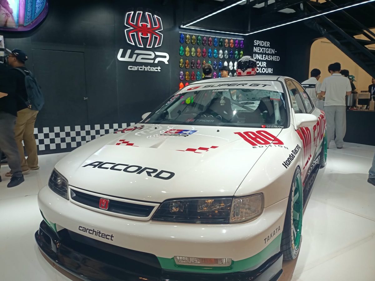 Sebuah Honda Accord bergaya racing dengan balutan warna putih, merah, dan hijau yang tampil mencolok di sebuah pameran otomotif. Mobil ini dilengkapi dengan body kit sporty, velg racing berwarna hijau, serta berbagai stiker khas motorsport. Latar belakangnya menampilkan booth modifikasi mobil dengan dekorasi bernuansa hitam dan logo Spider. Perpaduan desain klasik dan sentuhan modifikasi modern membuat mobil ini terlihat unik, agresif, dan penuh karakter.