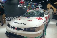 Sebuah Honda Accord bergaya racing dengan balutan warna putih, merah, dan hijau yang tampil mencolok di sebuah pameran otomotif. Mobil ini dilengkapi dengan body kit sporty, velg racing berwarna hijau, serta berbagai stiker khas motorsport. Latar belakangnya menampilkan booth modifikasi mobil dengan dekorasi bernuansa hitam dan logo Spider. Perpaduan desain klasik dan sentuhan modifikasi modern membuat mobil ini terlihat unik, agresif, dan penuh karakter.