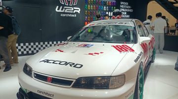 Sebuah Honda Accord bergaya racing dengan balutan warna putih, merah, dan hijau yang tampil mencolok di sebuah pameran otomotif. Mobil ini dilengkapi dengan body kit sporty, velg racing berwarna hijau, serta berbagai stiker khas motorsport. Latar belakangnya menampilkan booth modifikasi mobil dengan dekorasi bernuansa hitam dan logo Spider. Perpaduan desain klasik dan sentuhan modifikasi modern membuat mobil ini terlihat unik, agresif, dan penuh karakter.